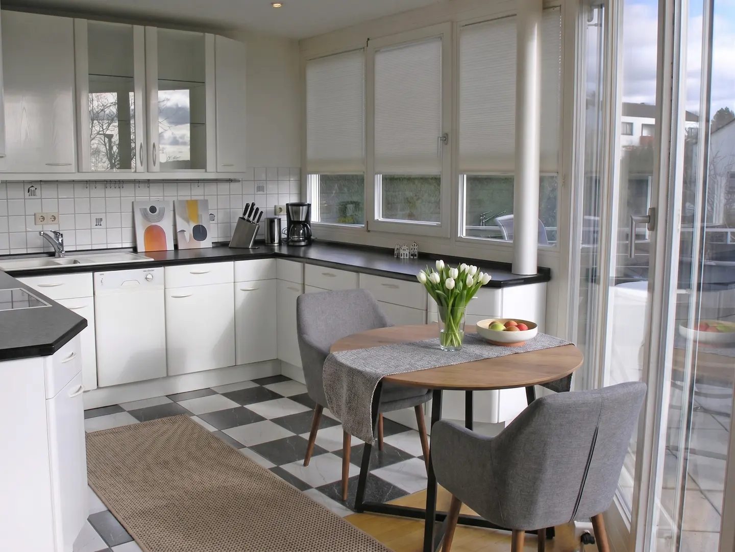 A bright kitchen with white cabinets, black countertops, and a black and white checkered floor. A round table with gray chairs sits near a glass door.