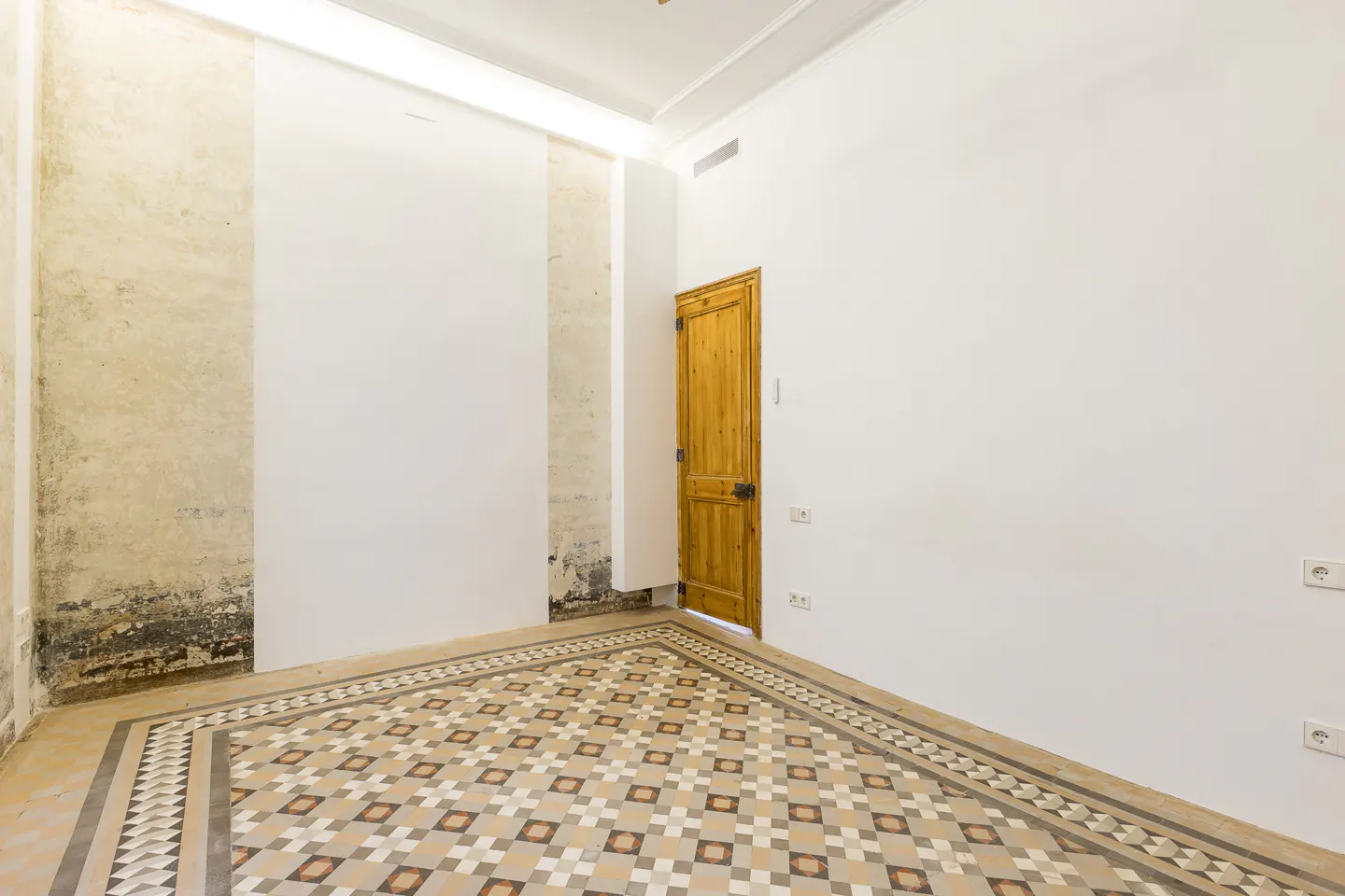 Empty room with patterned tile floor, white walls, and a wooden door slightly ajar. One wall has exposed plaster.