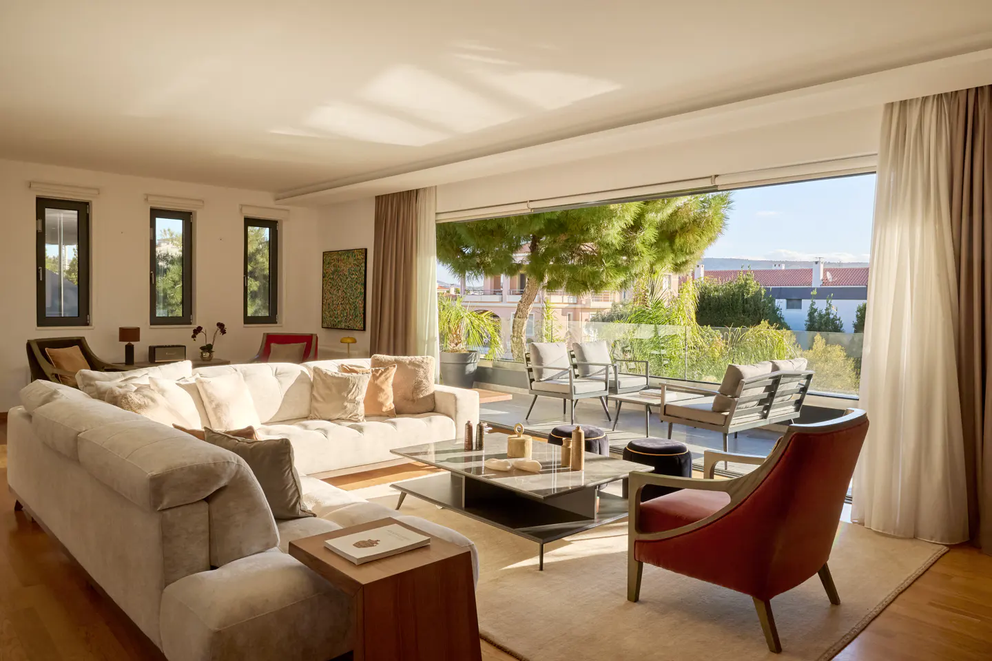 Bright living room with a large white sofa, a glass coffee table, and a red chair. A balcony with outdoor seating is visible through a large window.