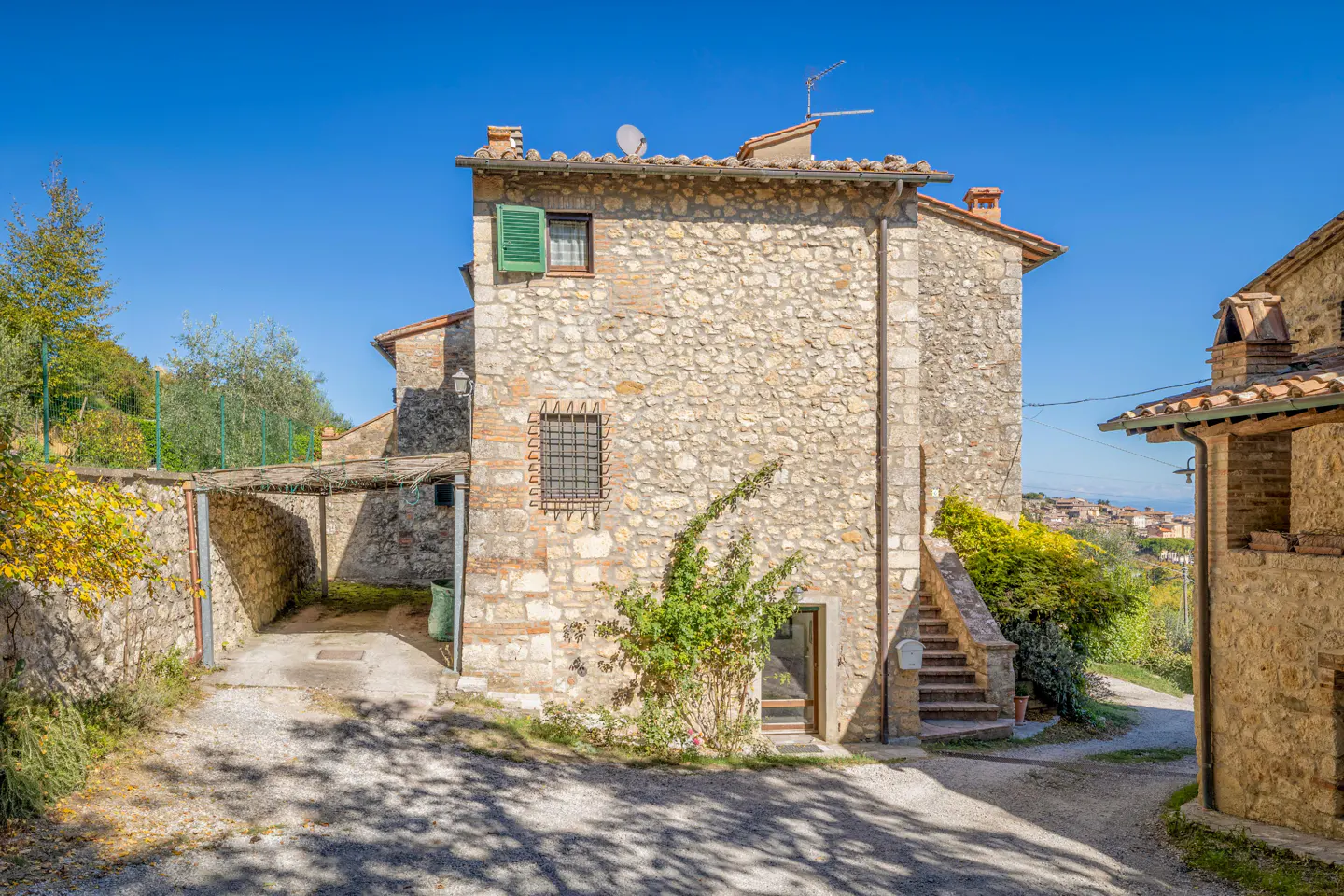 Exterior view of a stone house with green shutters and a red tile roof under a clear blue sky. A stone carport is attached to the side.