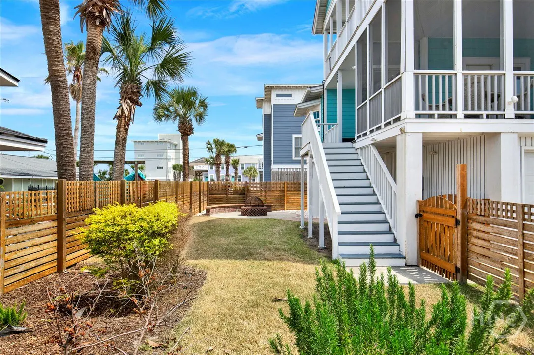 Backyard view of a beach house with a fire pit, wooden fence, palm trees, and white stairs leading to a screened porch.