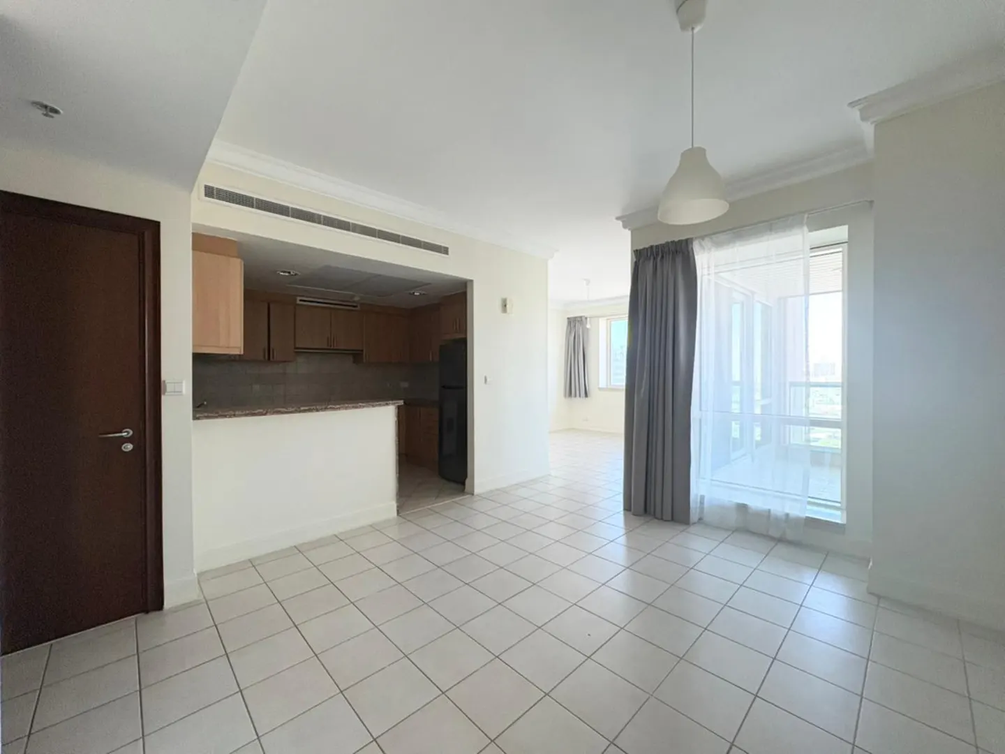 Bright, empty apartment with white tile floors. Kitchen with wood cabinets is visible. Large window with gray curtains lets in natural light.