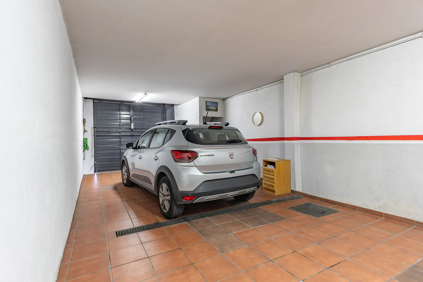 Indoor garage view with a silver car parked on brown tiled floor, white walls, and a closed gray metal door.