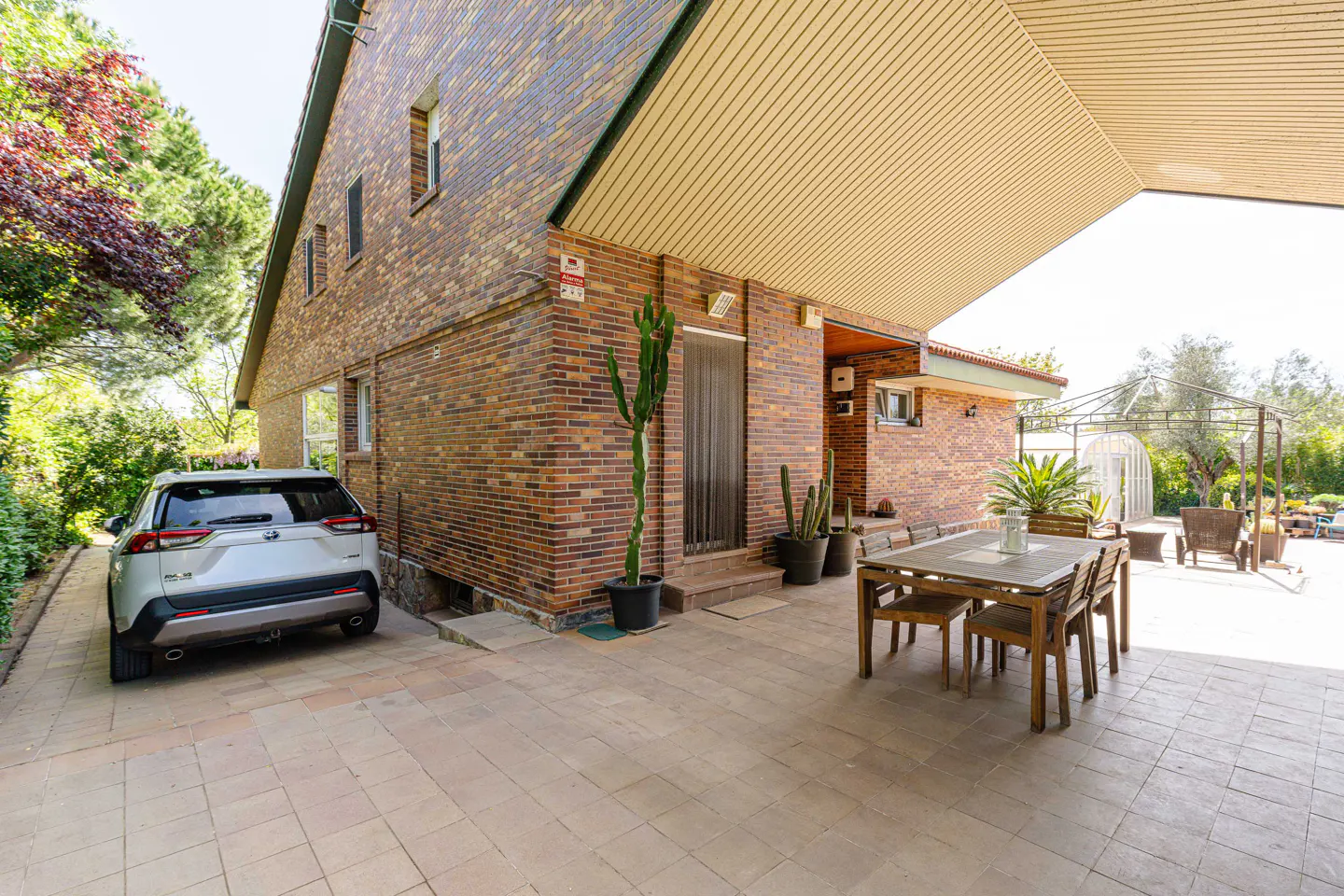 Brick house exterior with a covered patio, a wooden table and chairs, potted cacti, and a white car parked in the driveway.