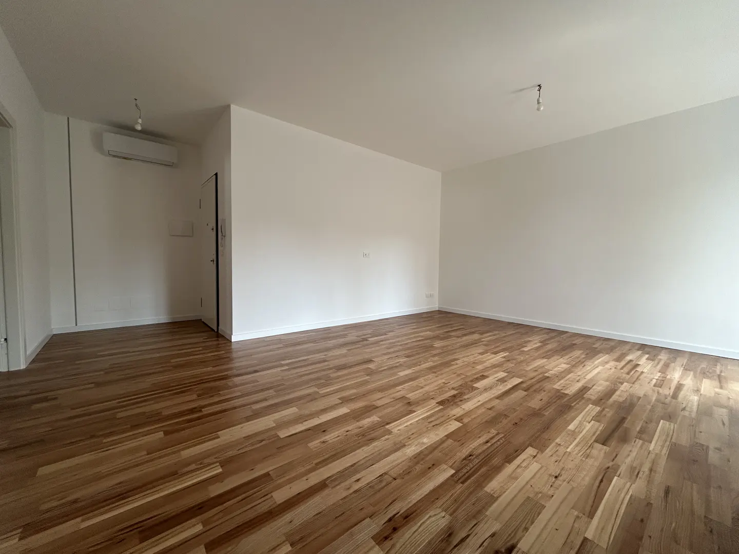 Empty room with light brown hardwood floors and white walls. A door and AC unit are visible.