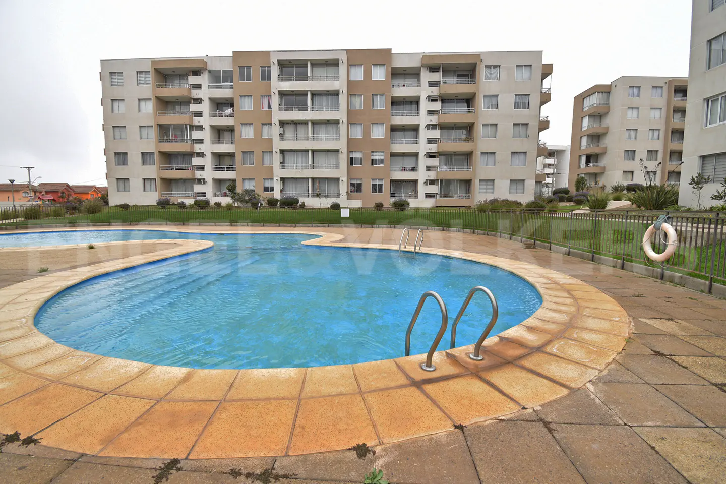 Outdoor pool with metal ladder and tan tile surround, in front of a multi-story apartment building. A life preserver hangs on a fence.
