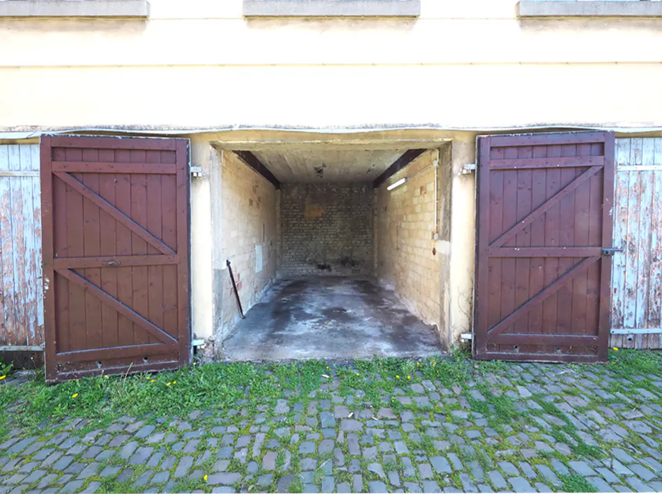 Open garage with brown wooden doors on a brick driveway. The garage interior is brick with a light fixture.