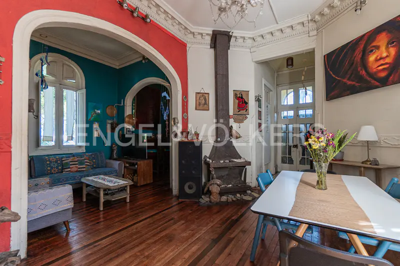 A living room with hardwood floors, a fireplace, and a dining table with blue chairs. Walls are red, teal, and white. A large painting hangs on the wall.
