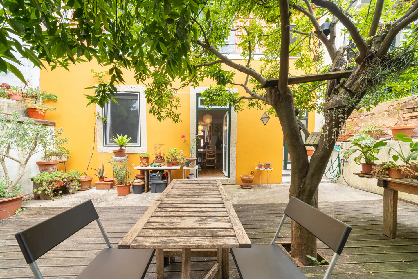 Outdoor patio with a wooden table and chairs, a yellow house with an open door, and potted plants.