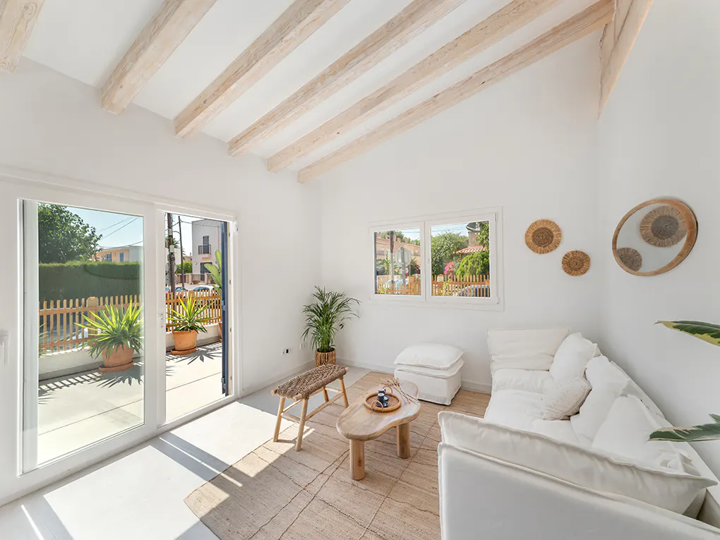 Bright living room with white walls, exposed beams, and a white sofa. A jute rug sits under a wooden coffee table and bench. Glass doors open to a patio.