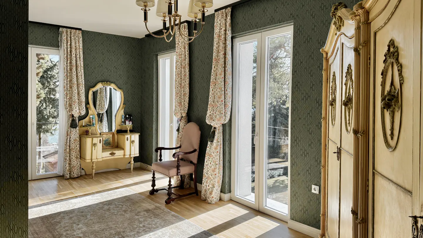 A bedroom with dark green walls, a light wood floor, and a large, ornate wardrobe. A vanity and chair sit near windows with floral curtains.