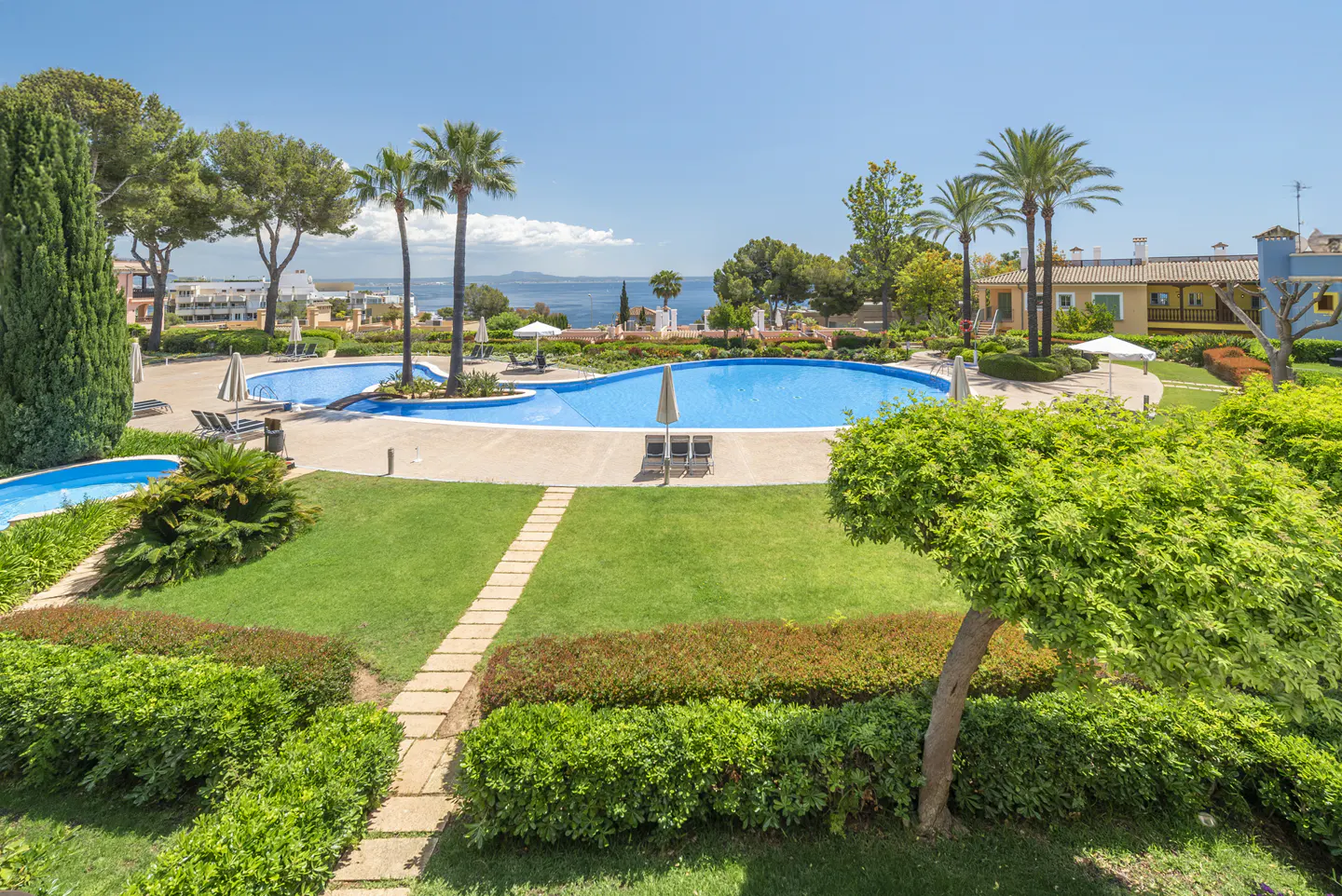 Aerial view of a resort with a blue pool, palm trees, green lawns, and a stone path leading through manicured hedges.