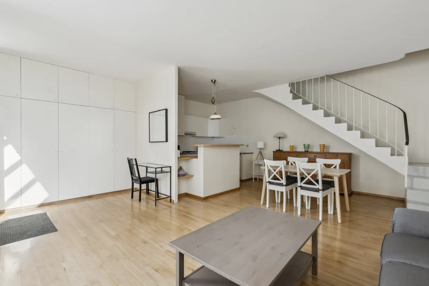 Bright, open-plan apartment with wooden floors, white walls, and a staircase. A dining table, kitchen, and built-in storage are visible.