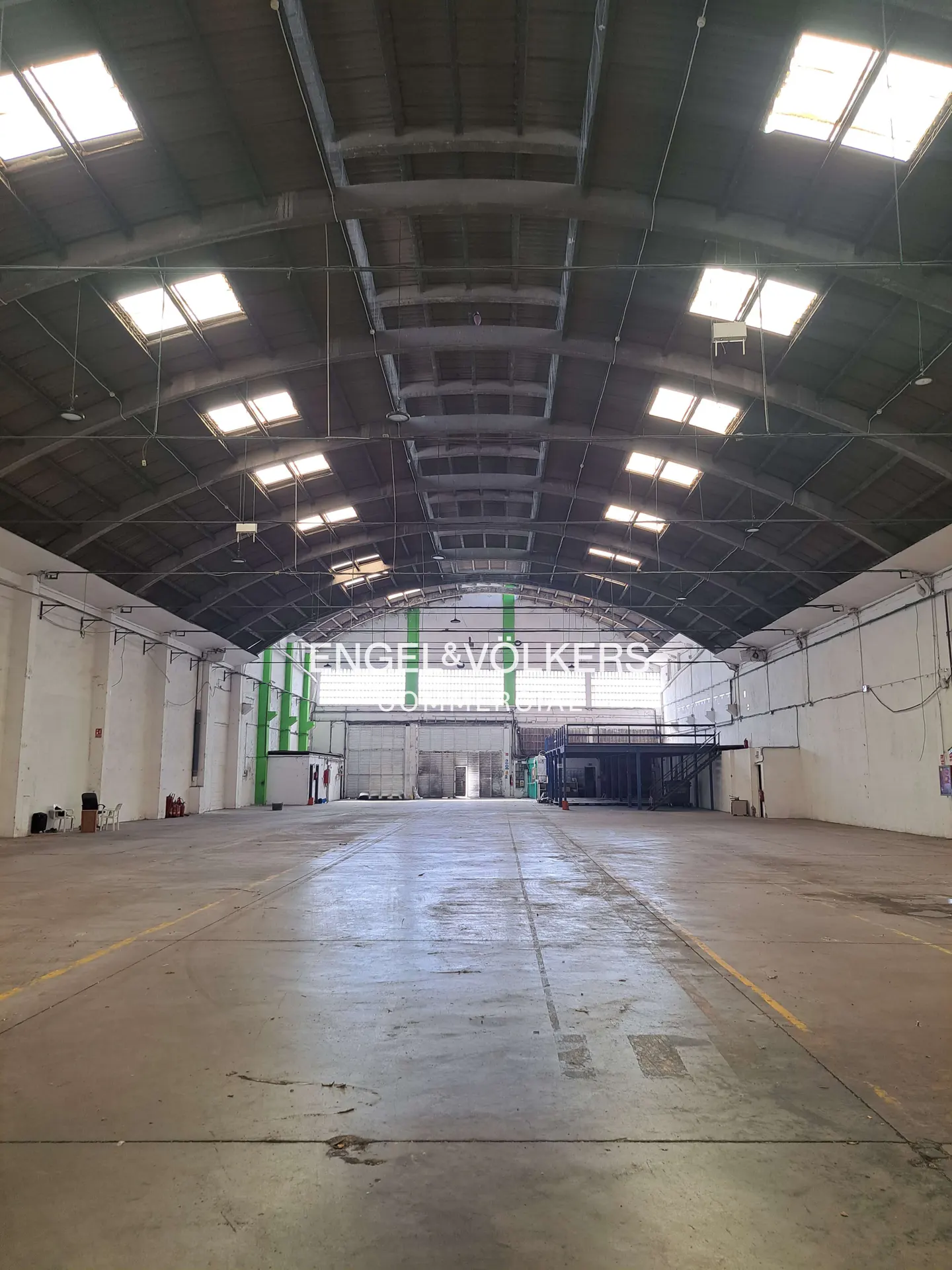 A wide shot of a large, empty warehouse with a concrete floor and a high, arched ceiling with skylights.