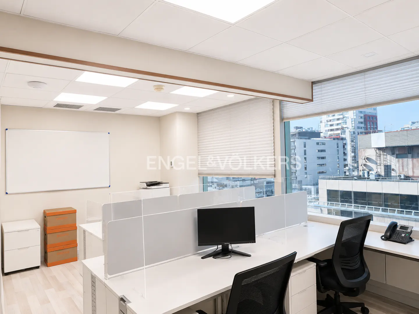 Bright office space with white desks, black chairs, and dividers. A whiteboard and city view are visible.