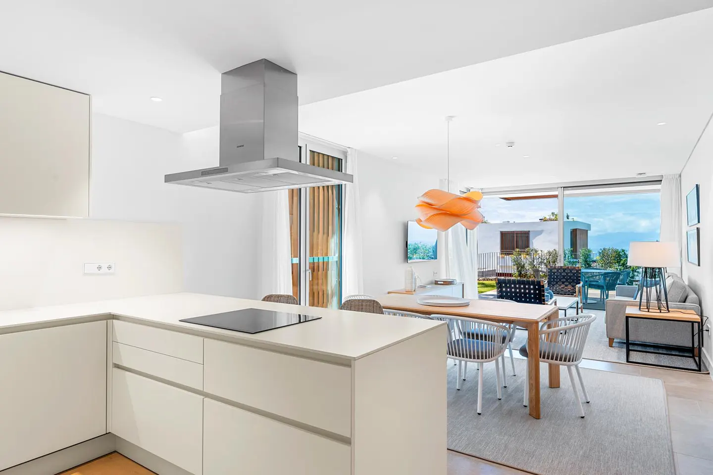 Bright, modern kitchen and dining area with white cabinets, stainless steel hood, and wood table with white chairs. Patio visible through sliding glass doors.