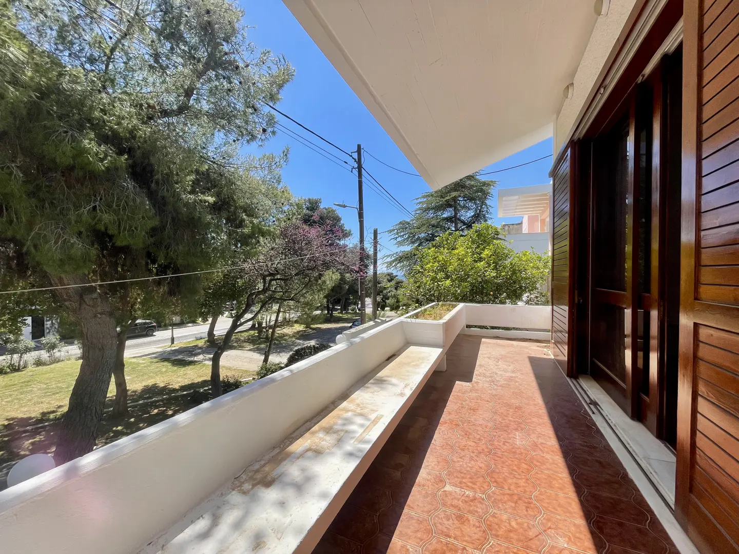 Balcony view with brown tile floor, white railing, and open wood-paneled doors. Trees and blue sky are visible in the background.