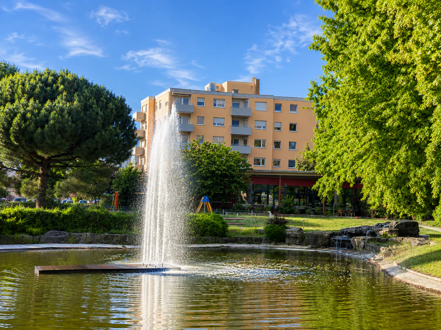 A fountain sprays water in a pond in front of a multi-story peach-colored apartment building surrounded by lush green trees.