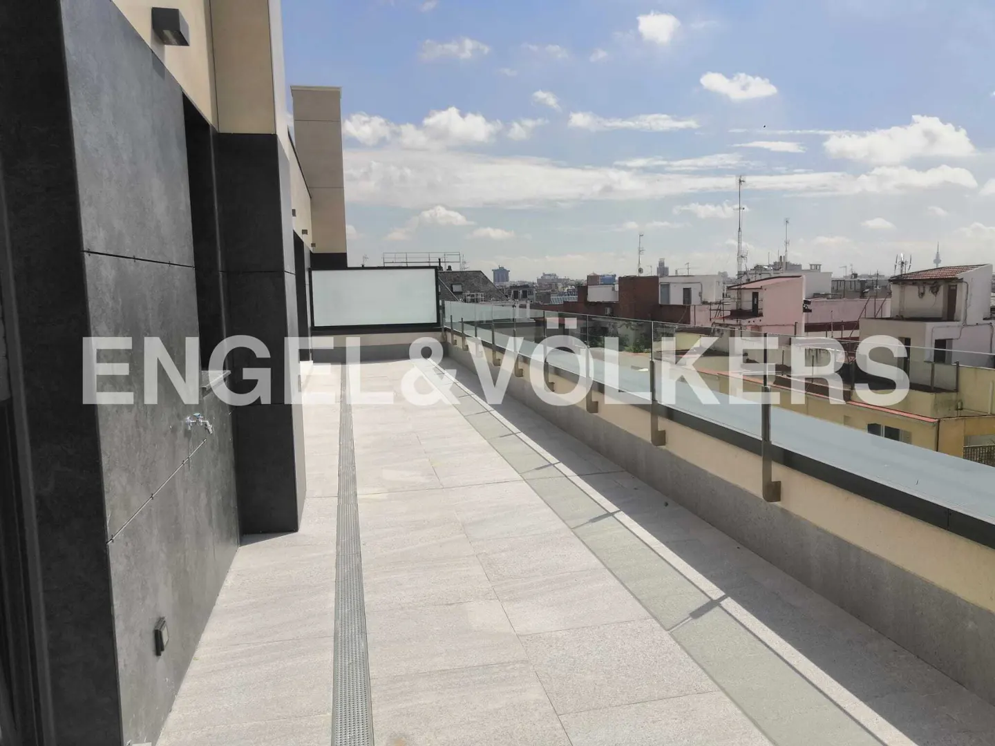 A rooftop terrace with a glass railing overlooks a city skyline under a cloudy sky.