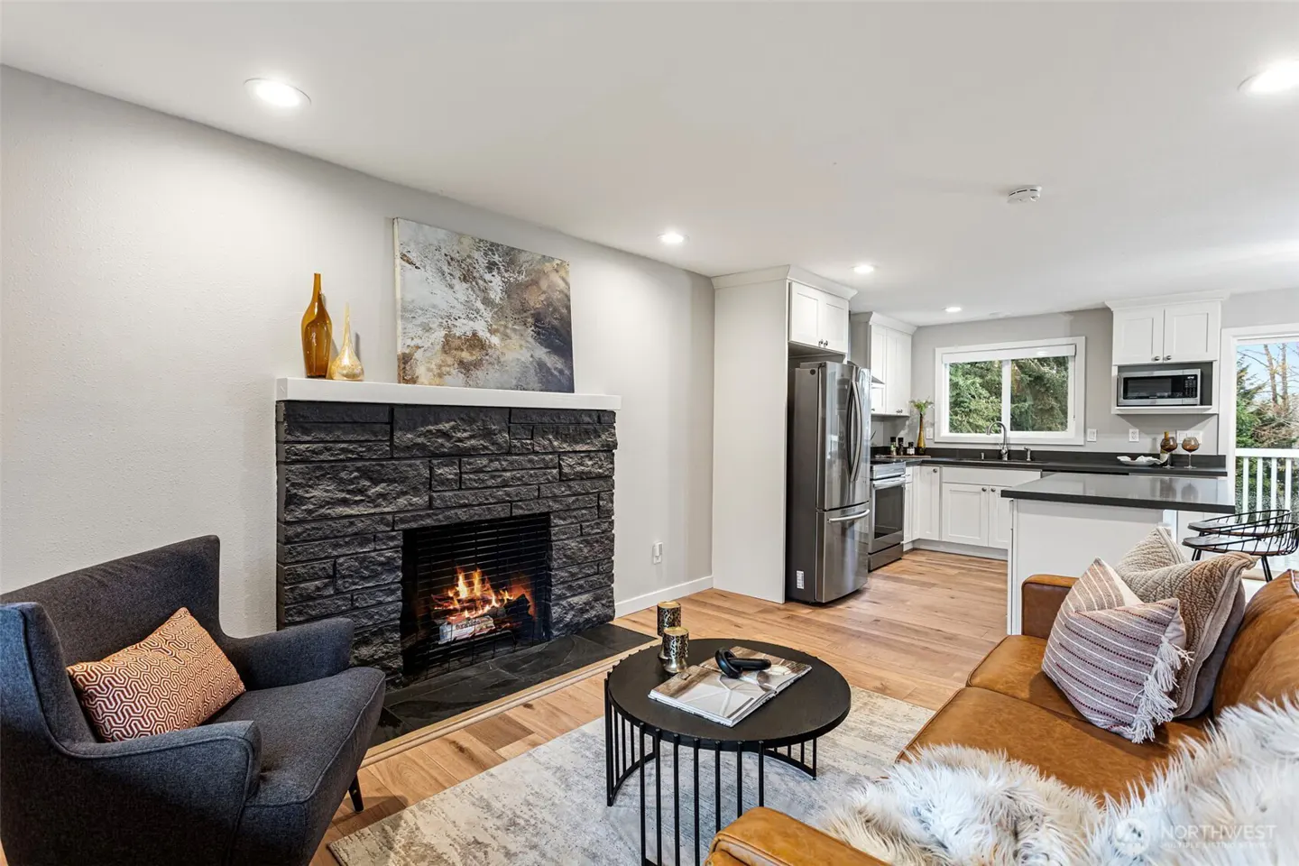 Modern living room with a black stone fireplace, gray armchair, tan leather sofa, and an open kitchen with white cabinets.