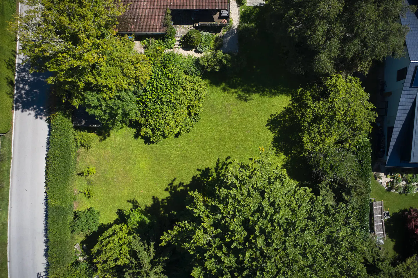 Aerial view of a rectangular green lawn surrounded by trees and hedges, next to a road and houses.