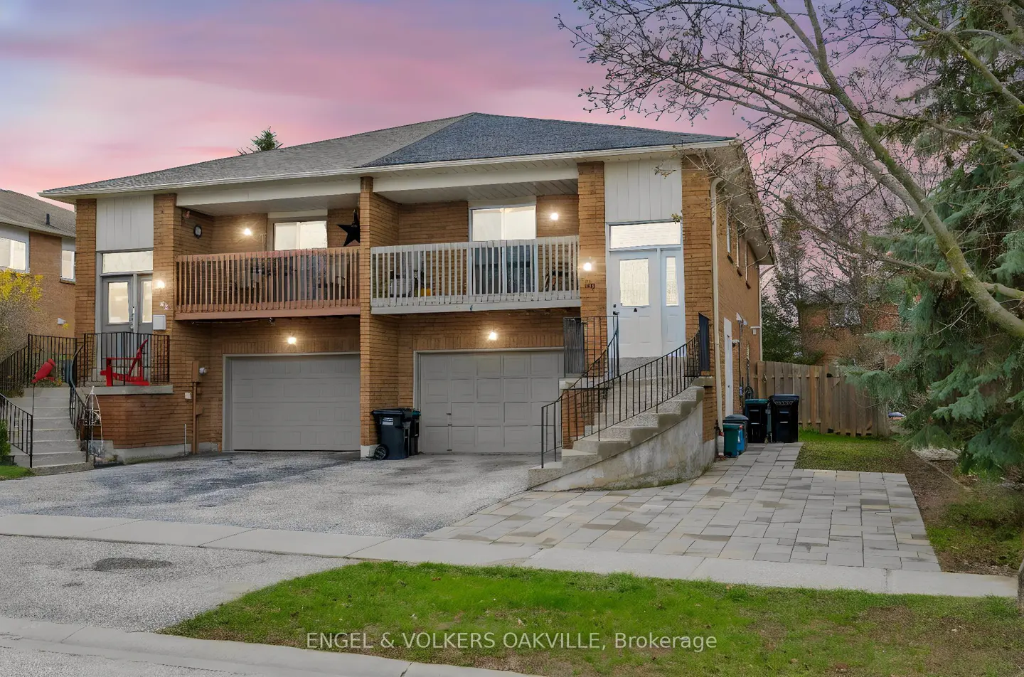 Exterior view of a two-story brick townhouse with balconies, gray garage doors, and a paved driveway. Dusk sky in the background.