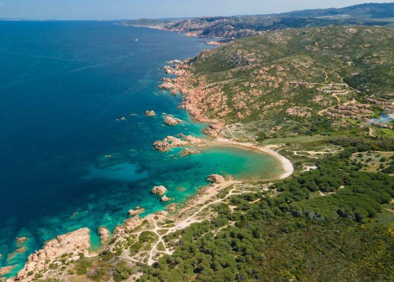 Aerial view of a turquoise Mediterranean cove with rocky cliffs, sandy beach, and lush green coastal vegetation.