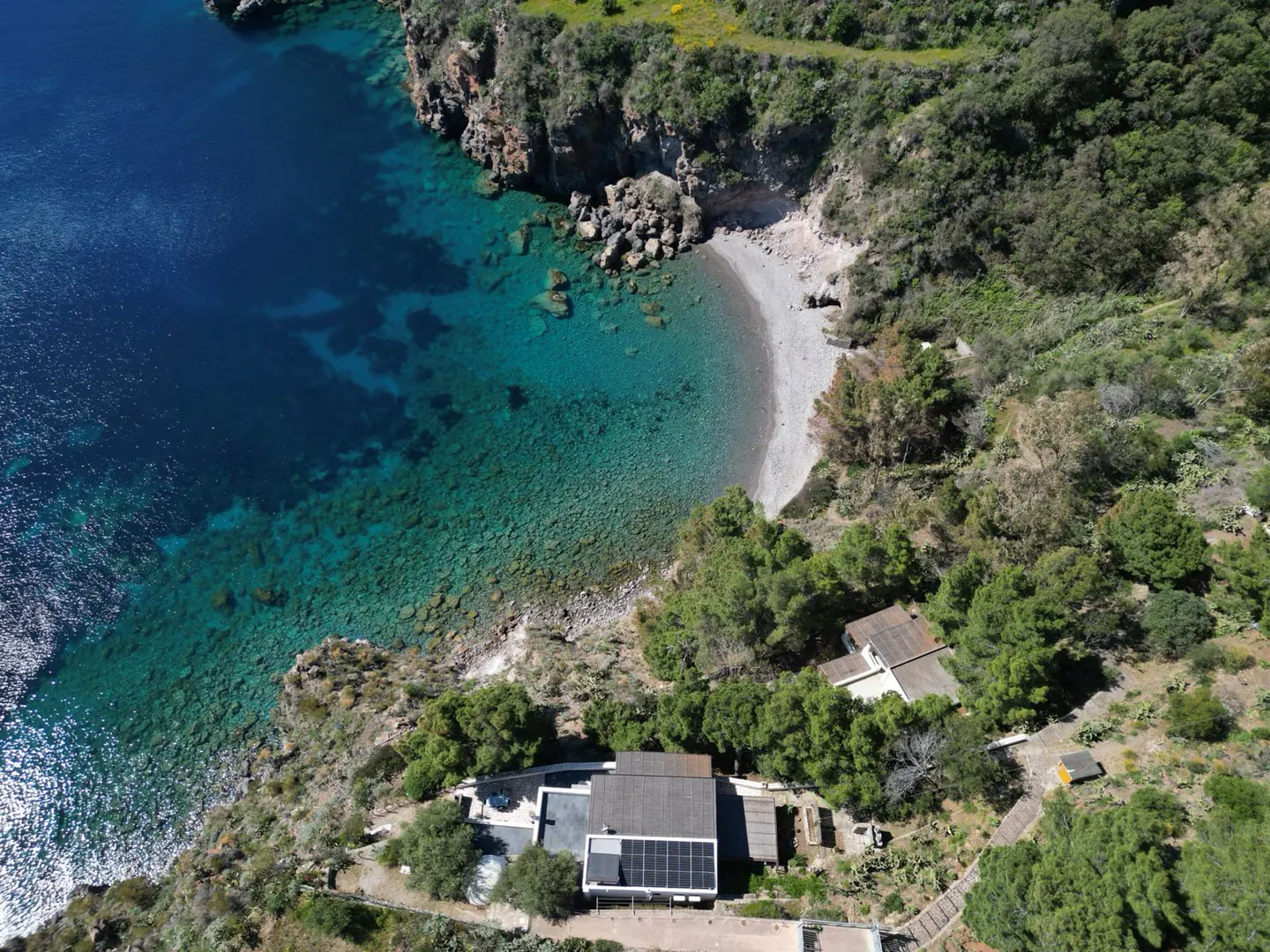 Aerial view of a coastal property with a house, solar panels, and a beach with turquoise water.