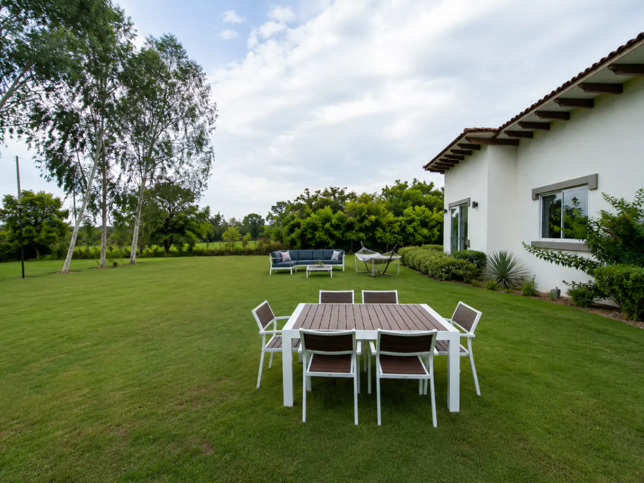 Outdoor patio with a white table and chairs on a green lawn, a white house with a brown roof in the background.