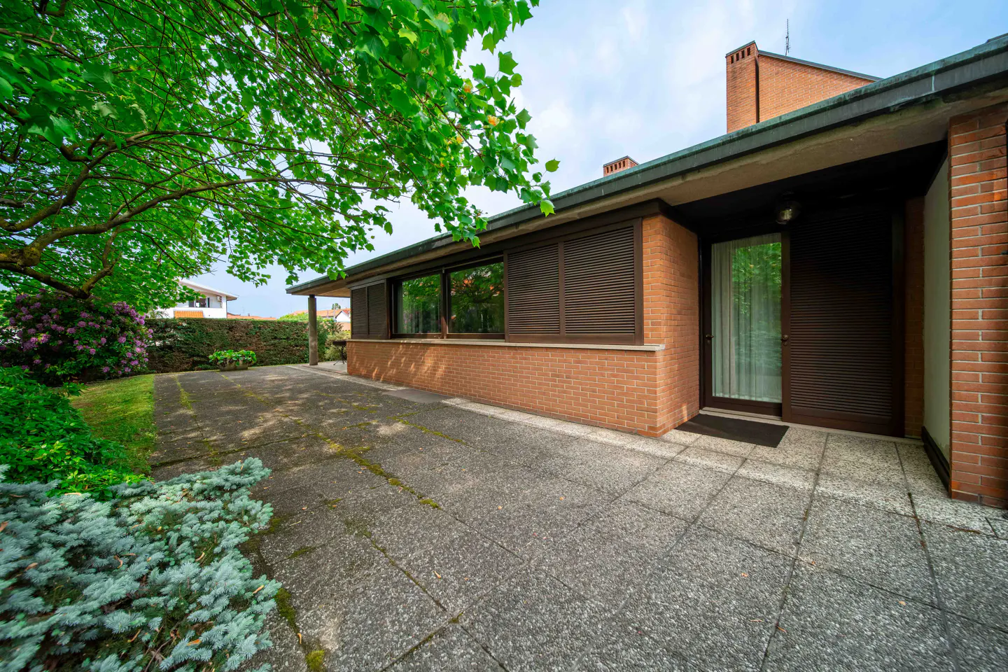 Exterior view of a one-story brick house with a concrete driveway and green foliage. The house has dark brown window shutters and a matching front door.