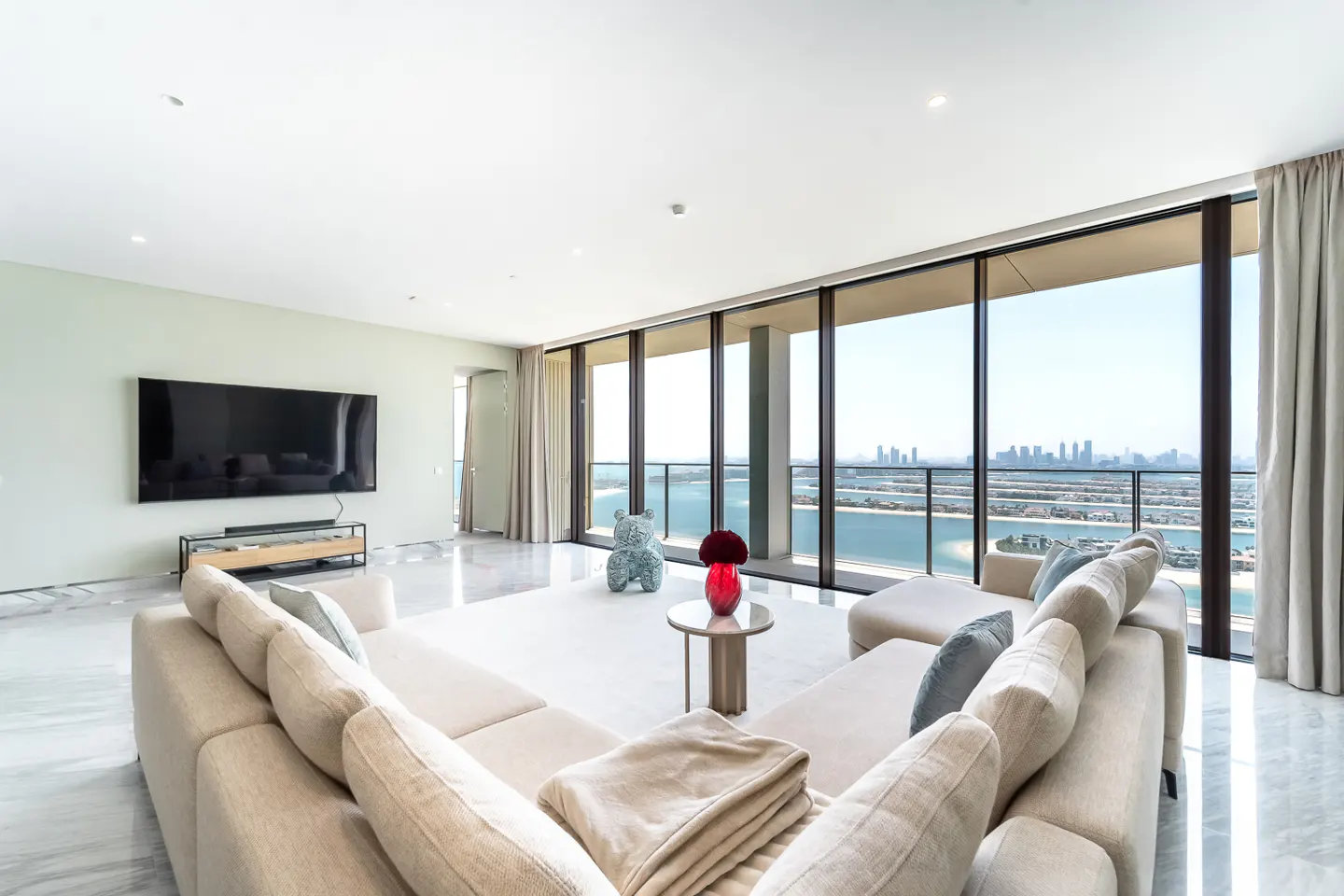 Bright living room with a beige sectional sofa, marble floors, and floor-to-ceiling windows with a city view. A large TV is mounted on the wall.