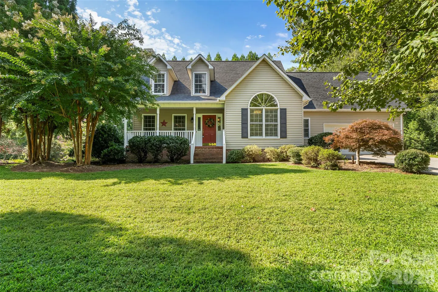 Beige two-story house with a red door, white porch, green lawn, and trees under a blue sky.