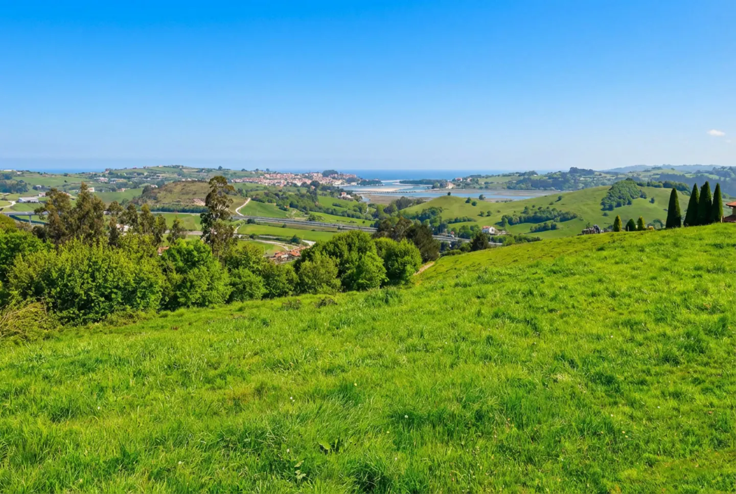 Scenic view of green hills under a blue sky, with a town and ocean in the distance.