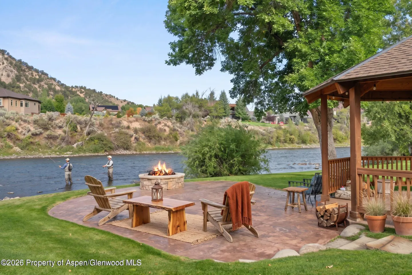 Outdoor patio with a fire pit, wooden chairs, and a table, next to a river where two people are fishing.