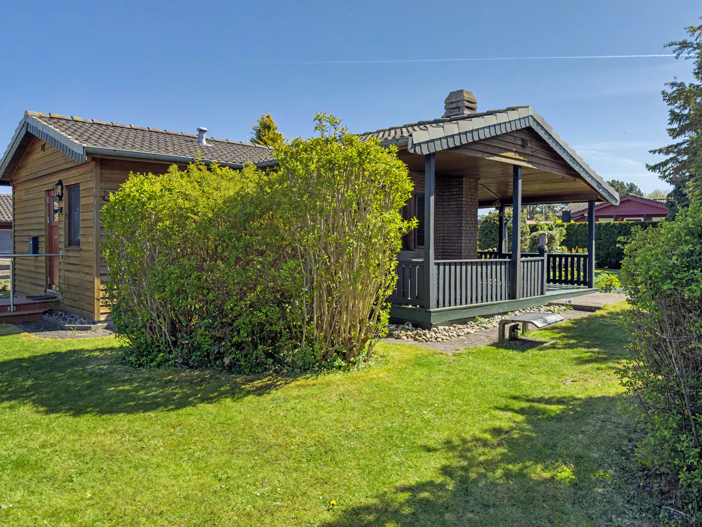 A wooden cabin with a covered porch, dark railings, and a green lawn under a clear blue sky.