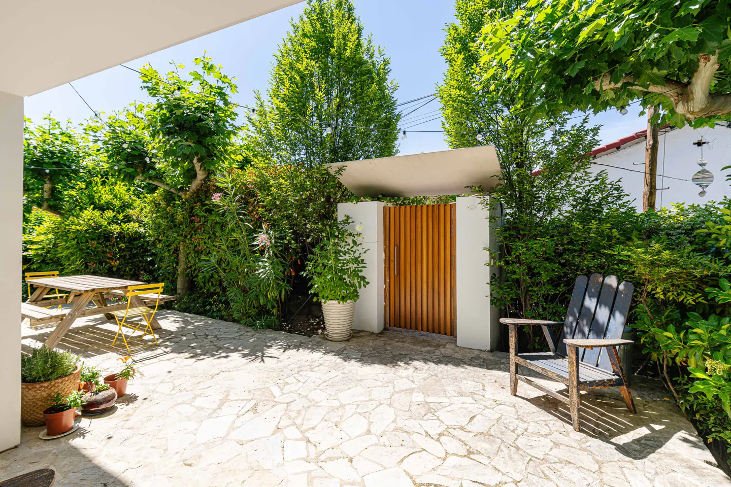 Outdoor patio with stone flooring, lush greenery, and a wooden gate. A picnic table and chair offer seating.