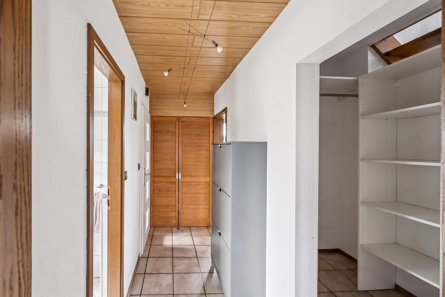 Hallway view with wood ceiling, tile floor, and white walls. A gray cabinet and white shelving unit are visible.