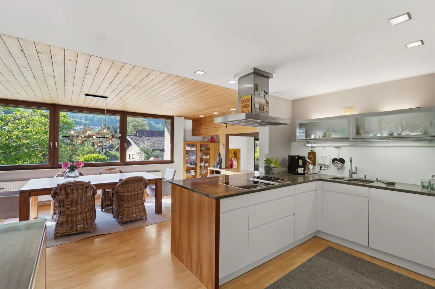 Modern kitchen with white cabinets, wood island, and dining area featuring wicker chairs under a wood-paneled ceiling.