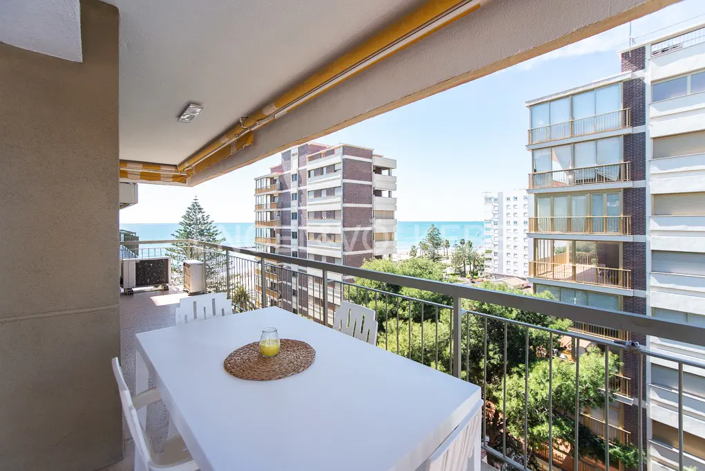 Balcony view with white table, chairs, and drink overlooking buildings, trees, and ocean on a sunny day.