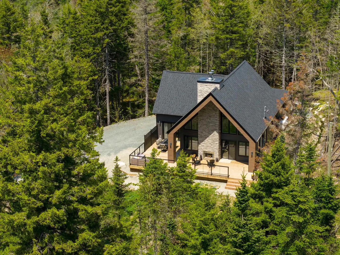 Aerial view of a modern dark-sided home with a stone chimney and large deck, nestled in a dense evergreen forest.