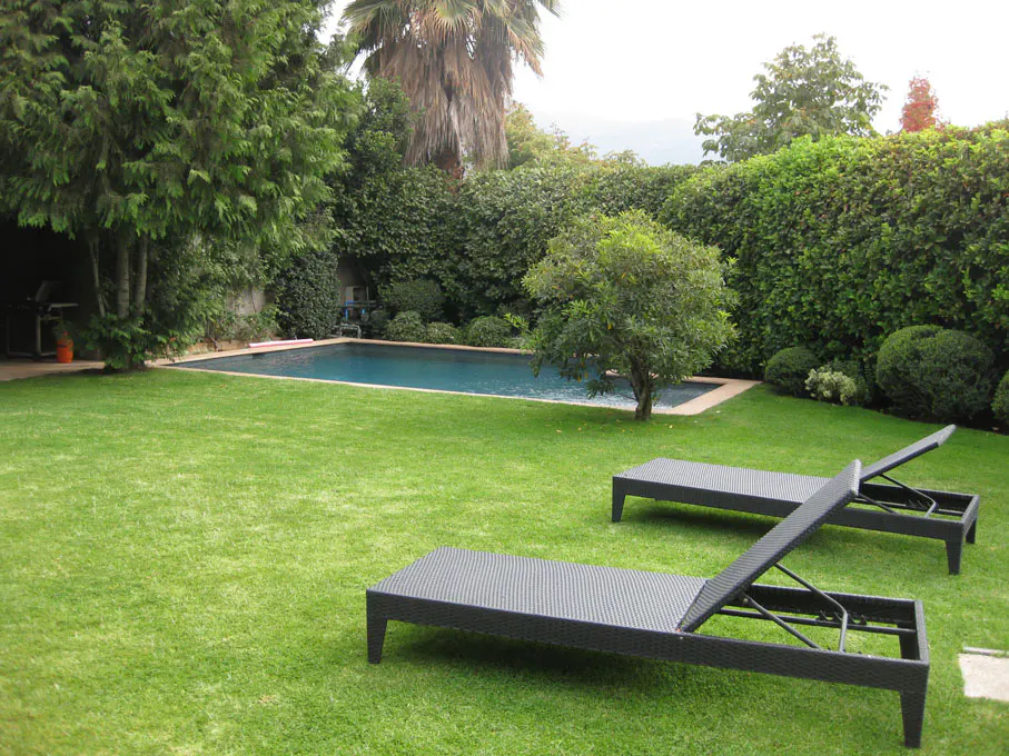 A backyard with a pool, green grass, trees, and two black lounge chairs.