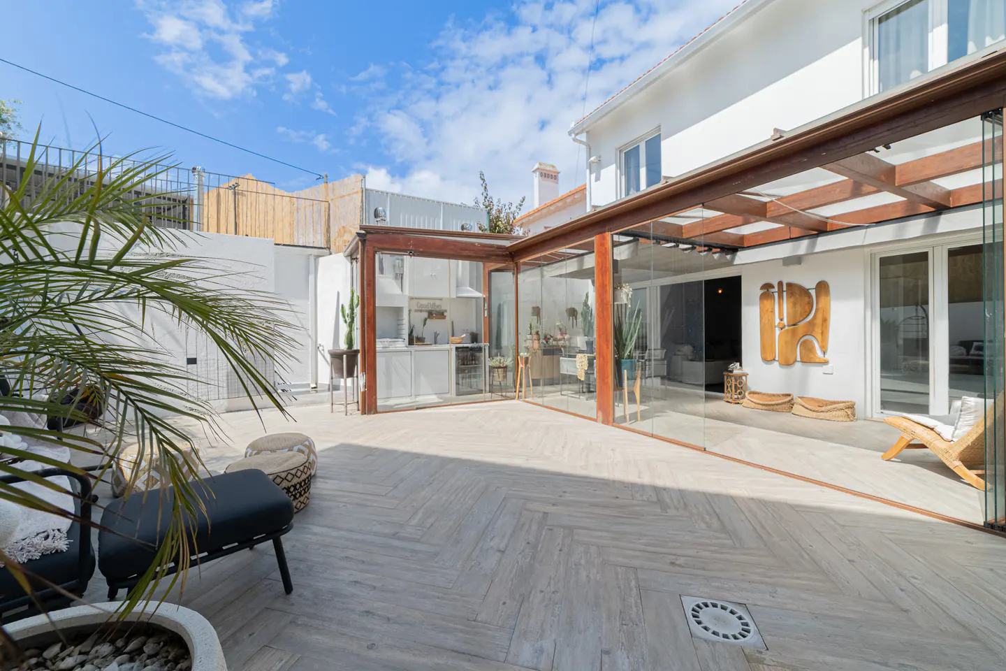 Outdoor patio with a glass-enclosed kitchen and seating area. The patio has light-colored herringbone tile and a wooden pergola.