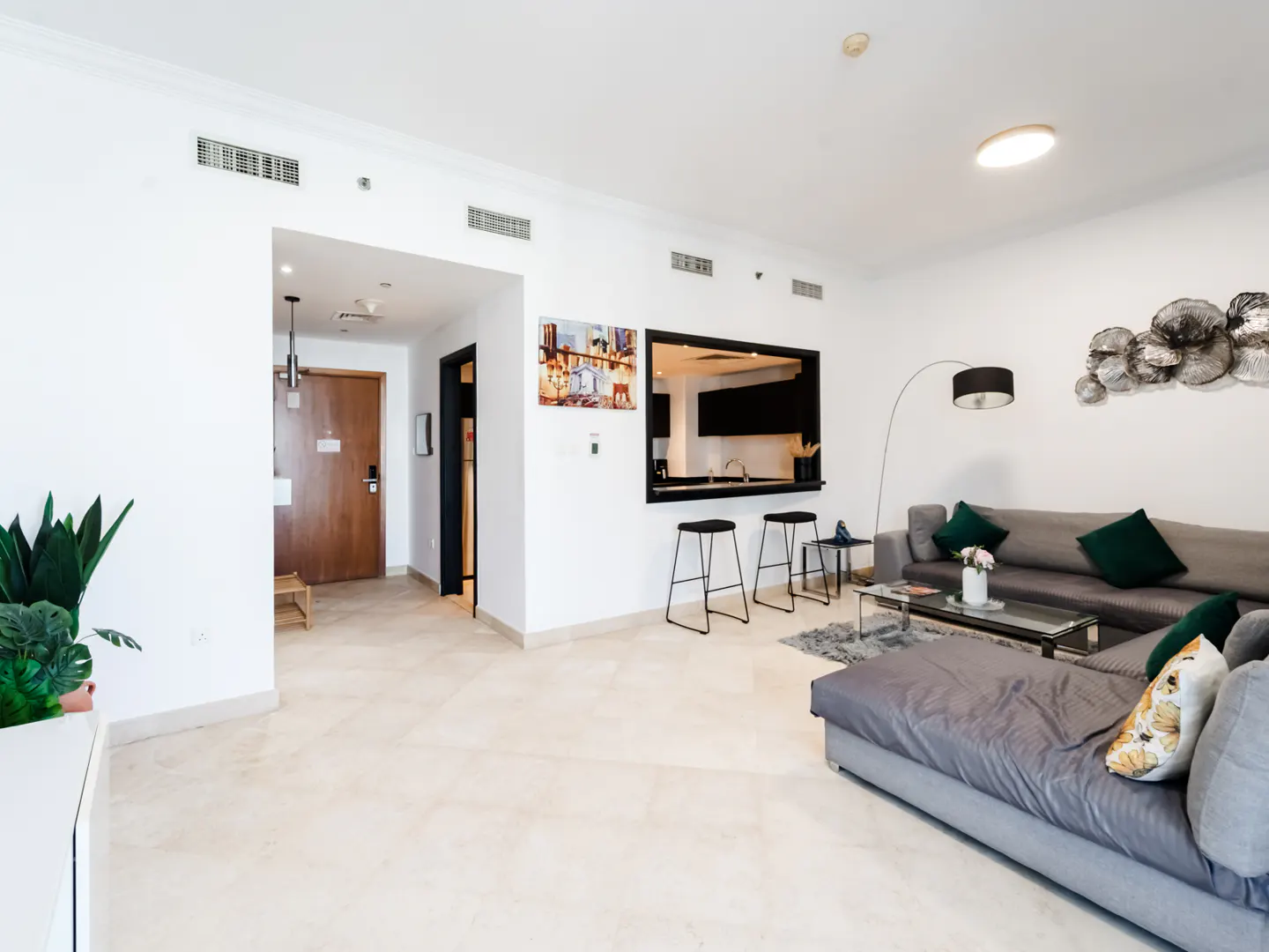 Modern living room with a grey sectional sofa, glass coffee table, breakfast bar with stools, and white tiled floors.