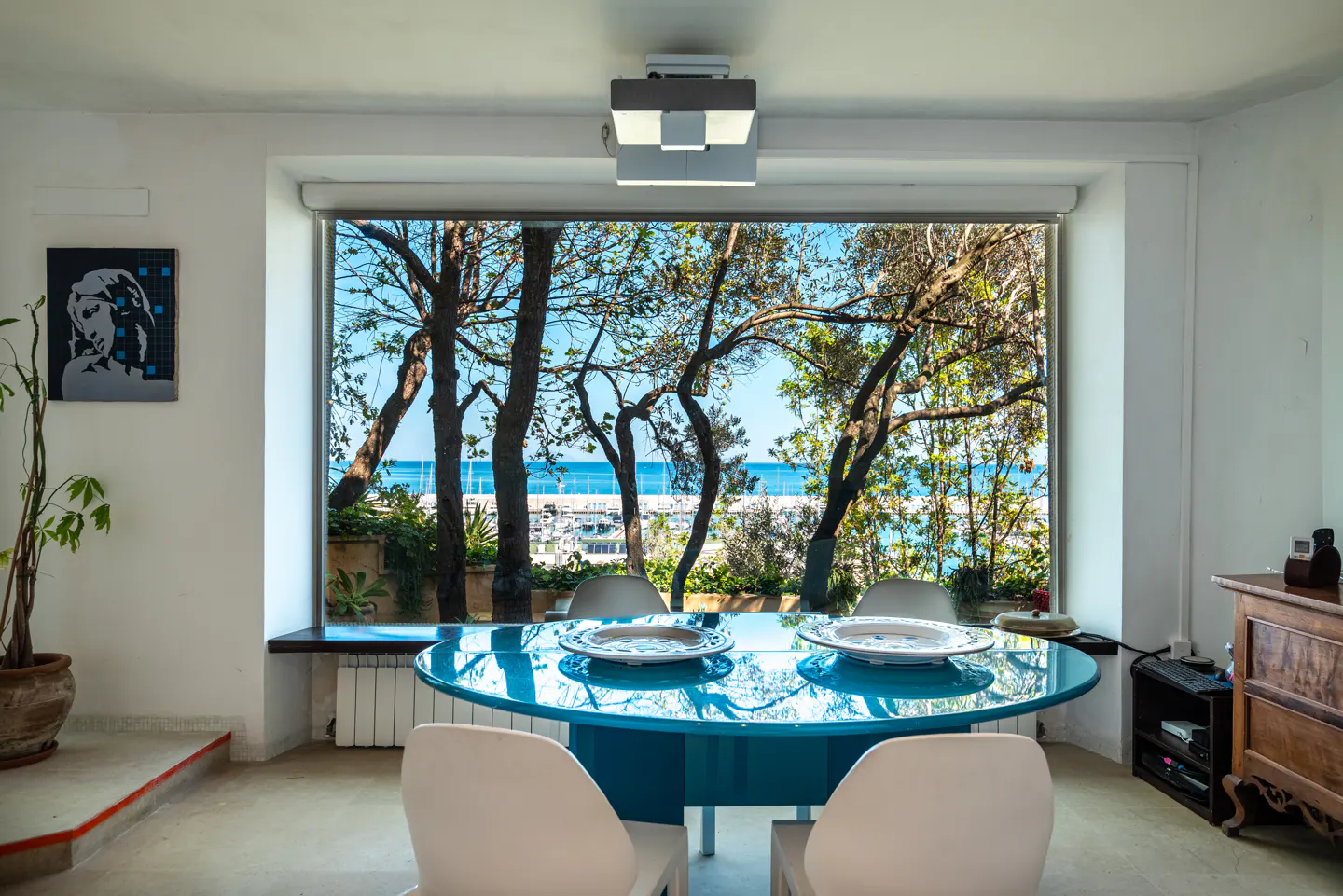 Bright dining room with a blue table and white chairs. A large window overlooks trees, the ocean, and a marina. Artwork hangs on the white wall.