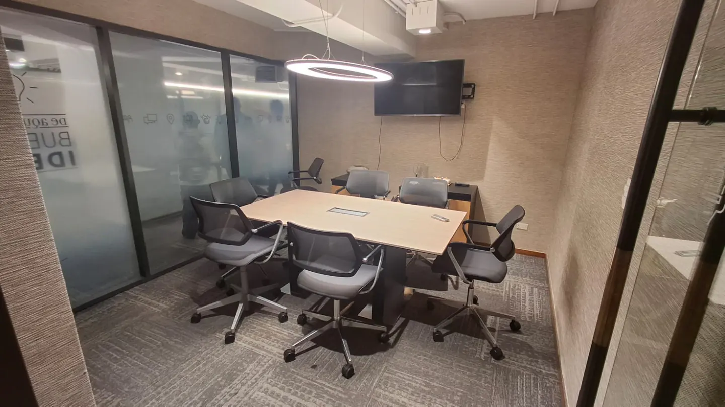 A small conference room with a light wood table, gray chairs, and a wall-mounted TV.