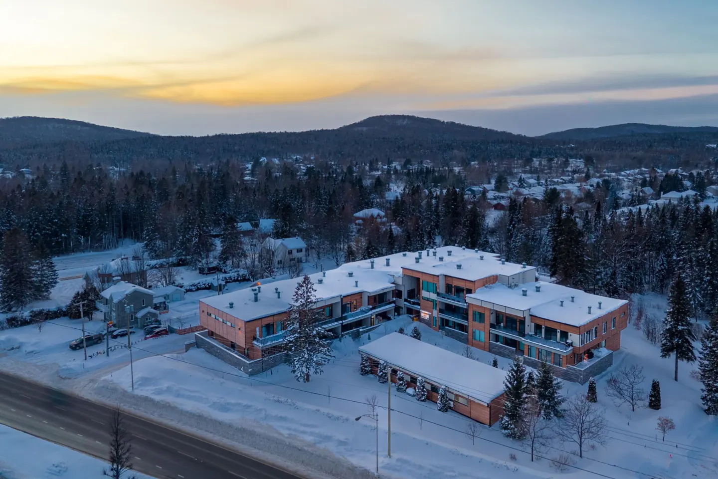 Aerial view of a modern apartment building with snow-covered roofs, surrounded by a forest and mountains at sunset.
