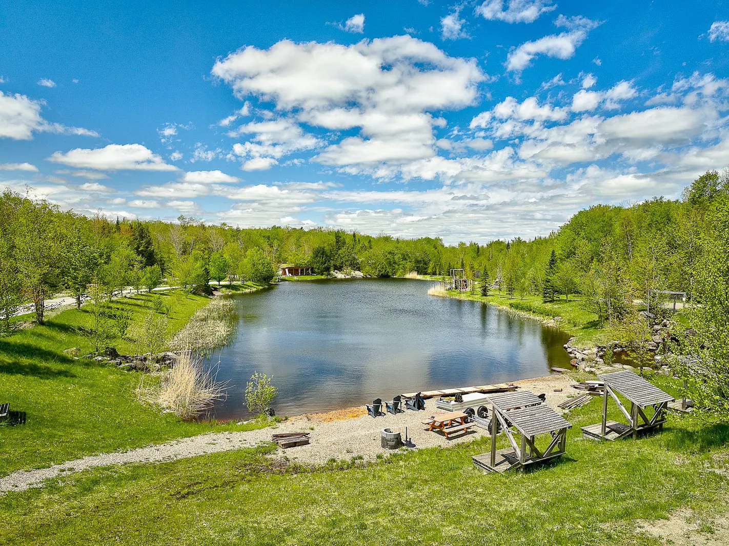 Scenic view of a lake surrounded by green trees under a blue sky with white clouds. A beach area has chairs, picnic table, and two small wooden structures.