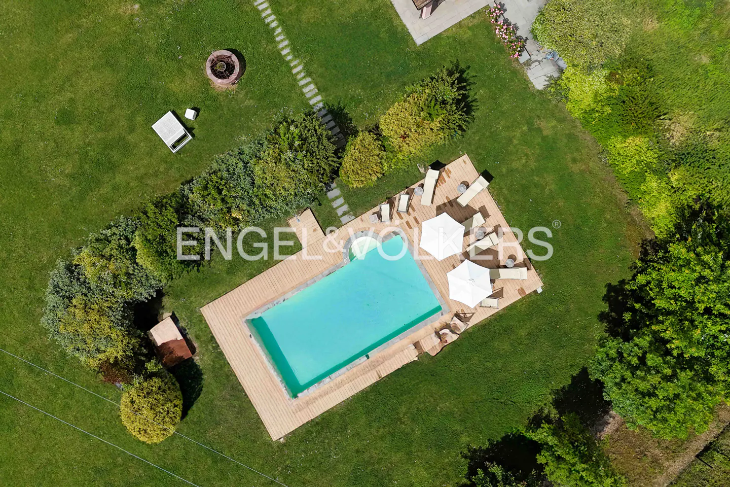 Aerial view of a rectangular pool with turquoise water, surrounded by a wooden deck, lounge chairs, and white umbrellas in a green lawn.
