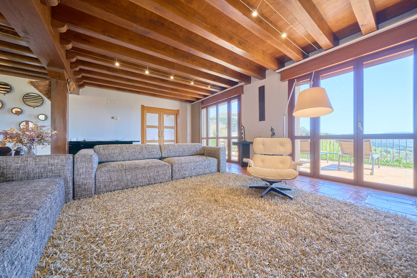 Living room with a gray sofa, cream chair, and a shaggy rug. The ceiling has wooden beams and there are large windows with a view.