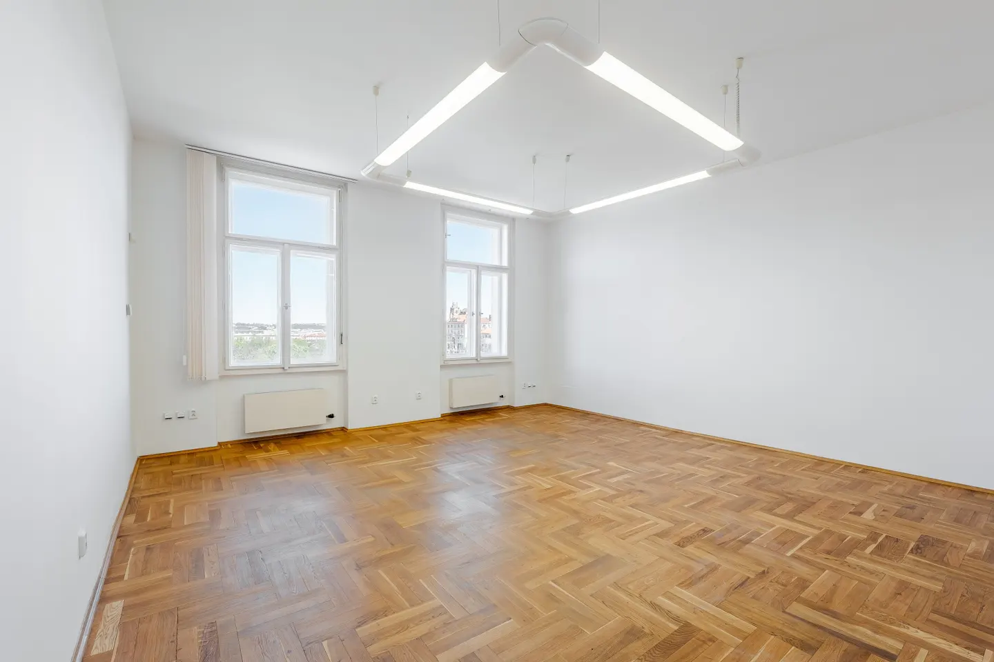 Bright, empty room with herringbone wood floors, white walls, two windows with city views, and a square fluorescent light fixture.